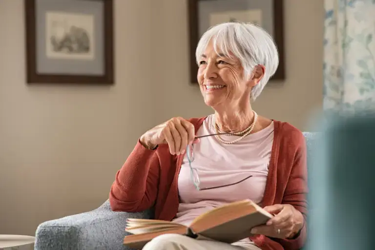 Dental Implants for Seniors Image - Elderly woman smiling on a sofa with a book on her hand